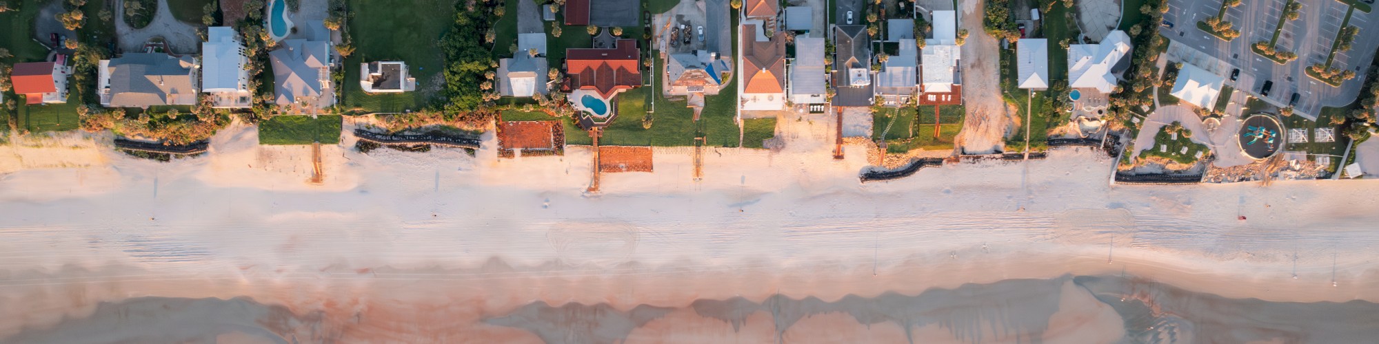 Aerial view of a beachfront neighborhood with houses, a road, sandy beach, and ocean waves hitting the shore.