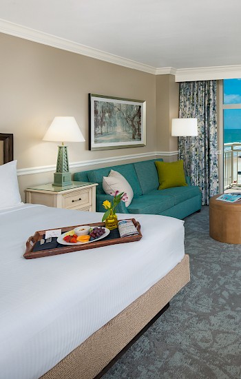 A hotel room with a bed, seating area, desk, and ocean view window. Decor is elegant with neutral tones and artistic accents.