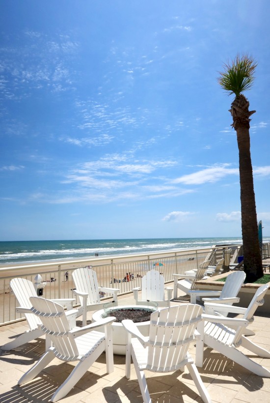 White Adirondack chairs encircle a fire pit on a beachfront patio, with a palm tree and ocean waves under a bright blue sky.