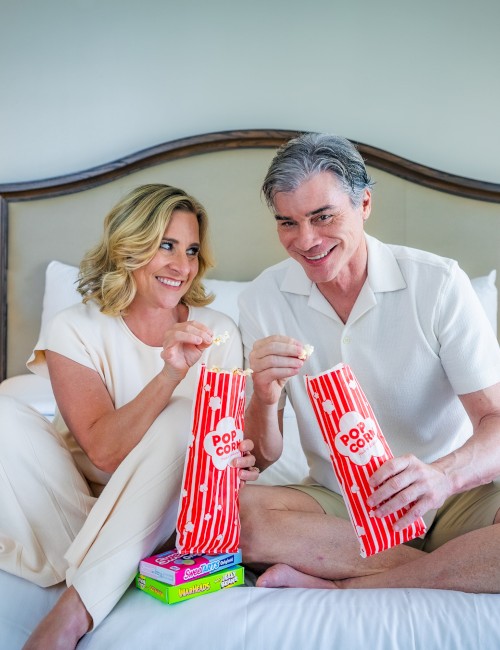 A couple sits on a bed, smiling and reaching into popcorn bags. They're enjoying snacks together in a cozy bedroom setting, with lamps on.