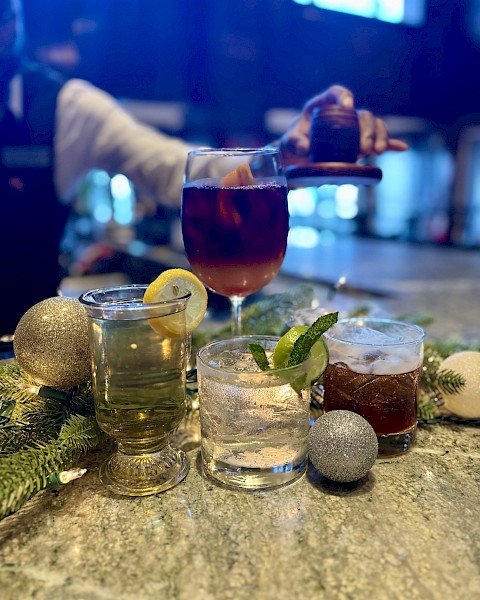 A selection of festive drinks with garnishes on a bar, surrounded by decorative ornaments and greenery.
