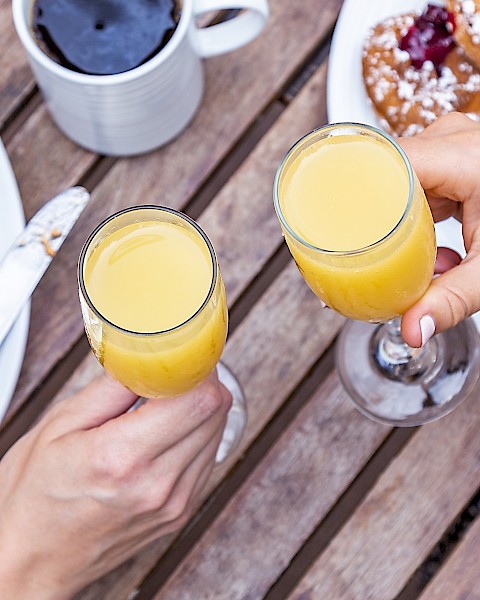 Two hands clinking glasses of orange juice, surrounded by breakfast foods and coffee on a wooden table.