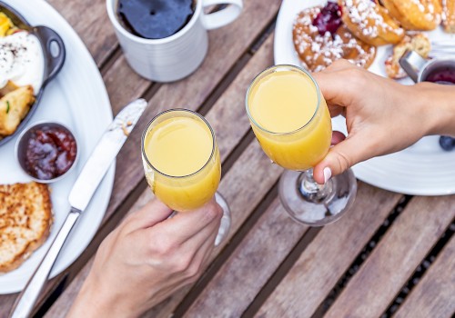 Two hands clinking glasses of orange juice, surrounded by breakfast foods and coffee on a wooden table.