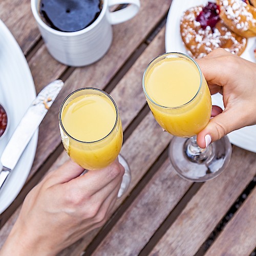 Two hands clinking glasses of orange juice, surrounded by breakfast foods and coffee on a wooden table.