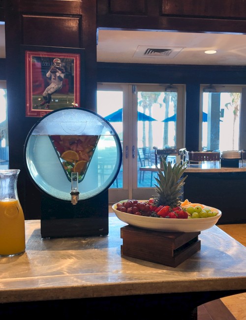 A restaurant with a drink station featuring juices, wine, and a fruit bowl. Large windows offer a view of an outdoor setting.