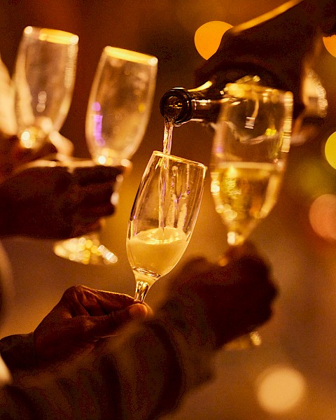 Four hands hold champagne glasses, with one being filled. The background features festive, blurred lights creating a celebratory ambiance.