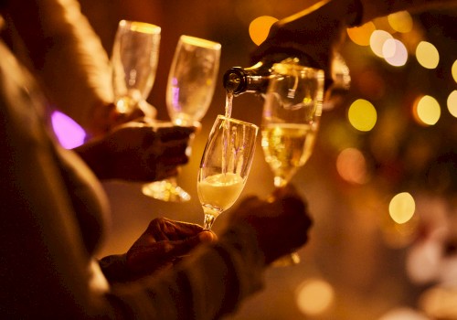 Four hands hold champagne glasses, with one being filled. The background features festive, blurred lights creating a celebratory ambiance.