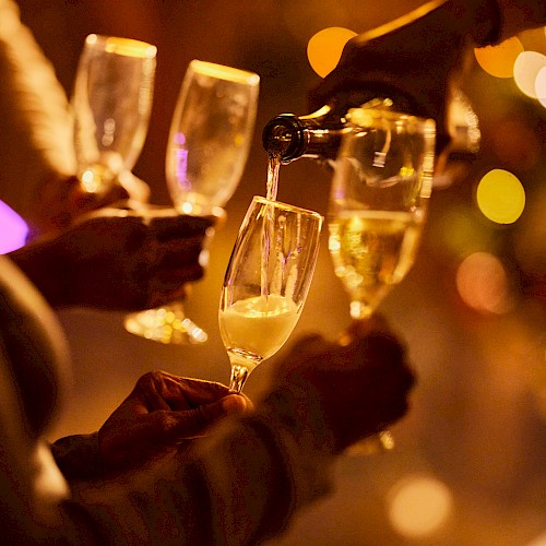 Four hands hold champagne glasses, with one being filled. The background features festive, blurred lights creating a celebratory ambiance.