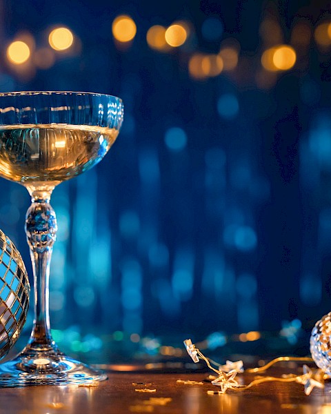 The image shows a champagne glass and disco balls on a festive table with blurred lights in the background, creating a party atmosphere.