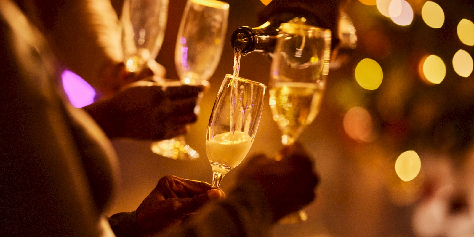 People are holding and pouring champagne into flutes, with festive, blurred lights in the background.