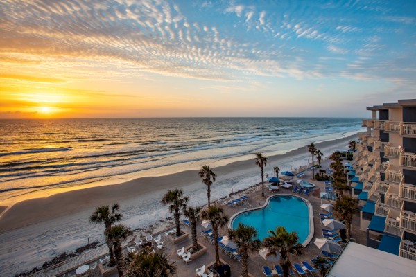 A beachfront view at sunset with a swimming pool, palm trees, beach chairs, and a hotel on the right side.