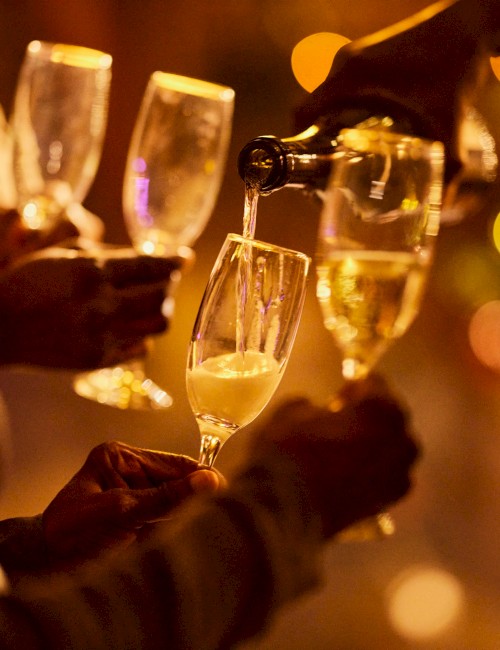 People are holding glasses as someone pours champagne, surrounded by warm, festive lights in the background.