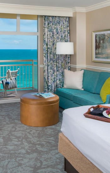 A hotel room with a bed, sofa, desk, and breakfast tray. A balcony overlooks the ocean with two chairs and a table, highlighted by sunlight.