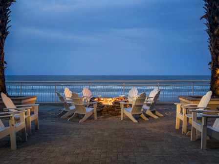 Chairs arranged around a fire pit with a view of the ocean. Two palm trees frame the scene under a blue evening sky.