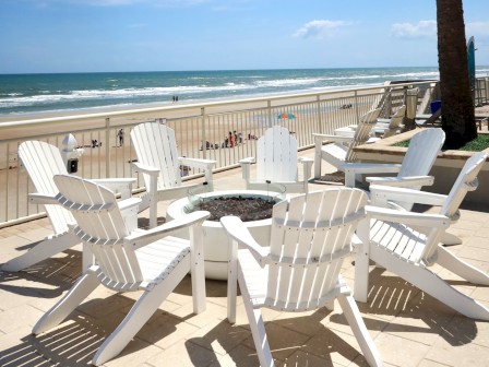 White chairs surround a fire pit on a sunny beachfront patio with a palm tree and ocean view.