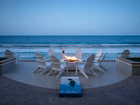 A serene seaside patio with a fire pit, Adirondack chairs, and a view of the ocean during evening.