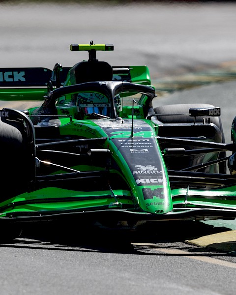 A green and black Formula 1 car is racing on a track, with various sponsor logos visible on its body, including “KICK”.