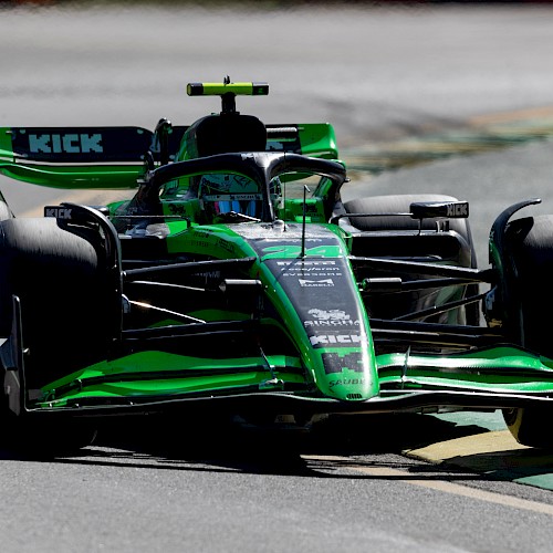 A green and black Formula 1 car is racing on a track, with various sponsor logos visible on its body, including “KICK”.