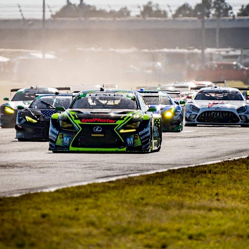 A group of race cars speeds down a track during a competition, with a green car leading the pack on a clear day.