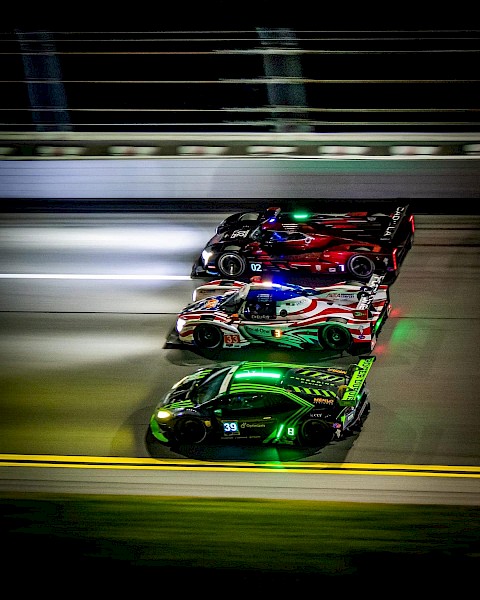 Three race cars are speeding side by side on a racetrack at night, with bright lights and motion blur capturing the sense of high speed.