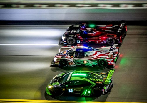 Three race cars are speeding side by side on a racetrack at night, with bright lights and motion blur capturing the sense of high speed.