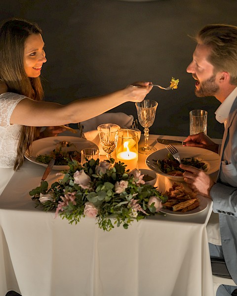 A couple enjoys a romantic candlelit dinner, with the woman feeding the man, at a beautifully decorated table with elegant settings.