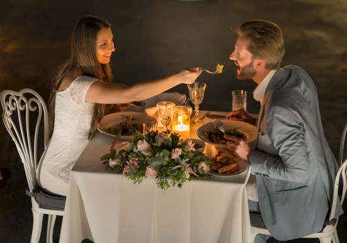 A couple enjoys a romantic candlelit dinner, with the woman feeding the man, at a beautifully decorated table with elegant settings.