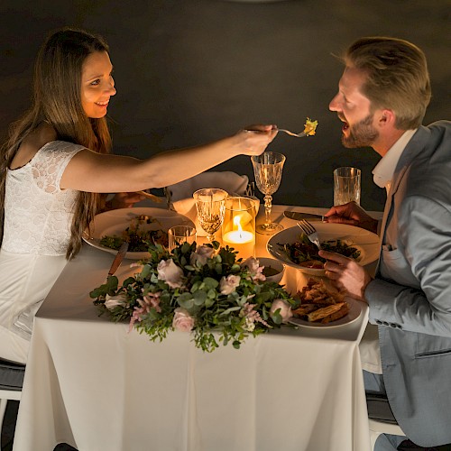 A couple enjoys a romantic candlelit dinner, with the woman feeding the man, at a beautifully decorated table with elegant settings.