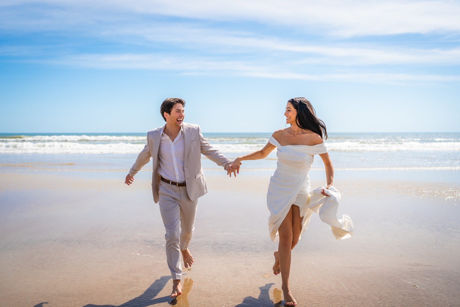 A couple joyfully runs barefoot on a beach, holding hands under a clear blue sky with ocean waves in the background.