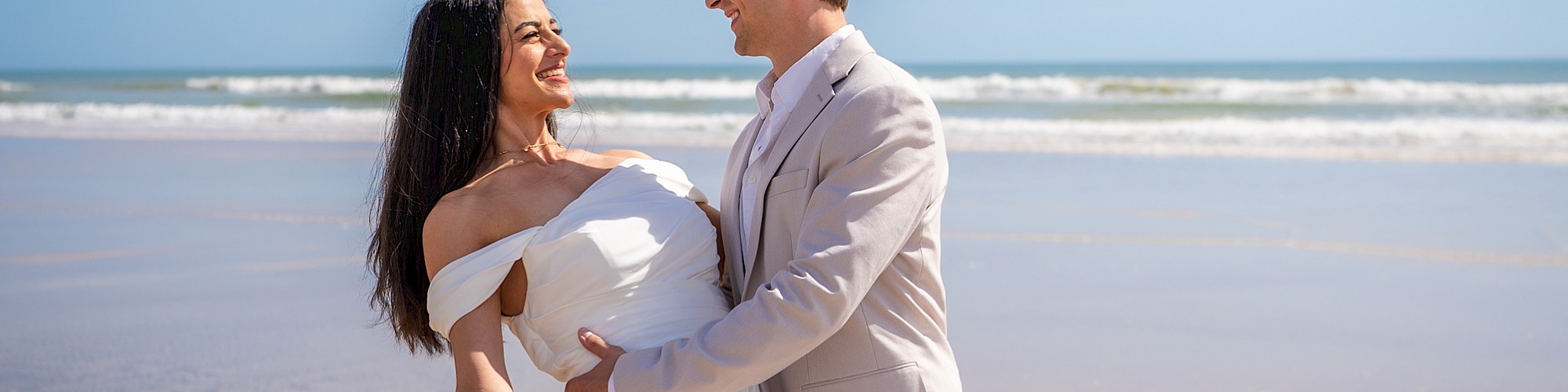 A couple in wedding attire is at the beach, with the groom holding the bride who holds a bouquet, against a blue sky and ocean backdrop.