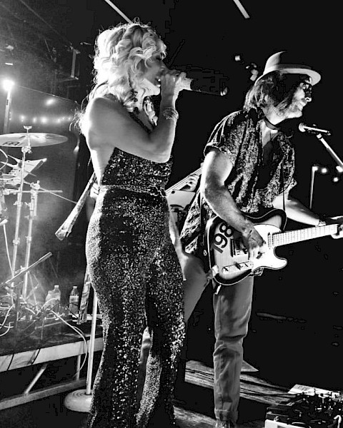 A black and white image of a band performing on stage. The singer holds a microphone and the guitarist plays, with drums visible in the background.