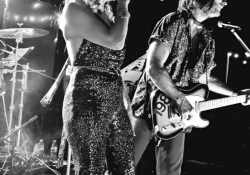 A black and white image of a band performing on stage. The singer holds a microphone and the guitarist plays, with drums visible in the background.