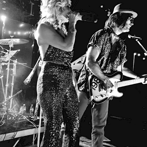 A black and white image of a band performing on stage. The singer holds a microphone and the guitarist plays, with drums visible in the background.