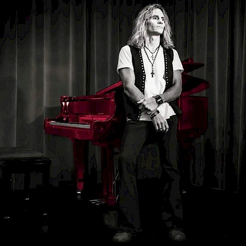 A person with long hair stands in front of a vibrant red piano on a dimly lit stage, with a curtain backdrop.