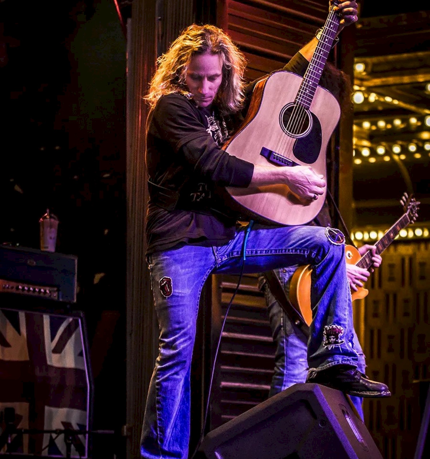 A musician with long hair energetically playing an acoustic guitar on stage, surrounded by vibrant lighting and a musical setup.