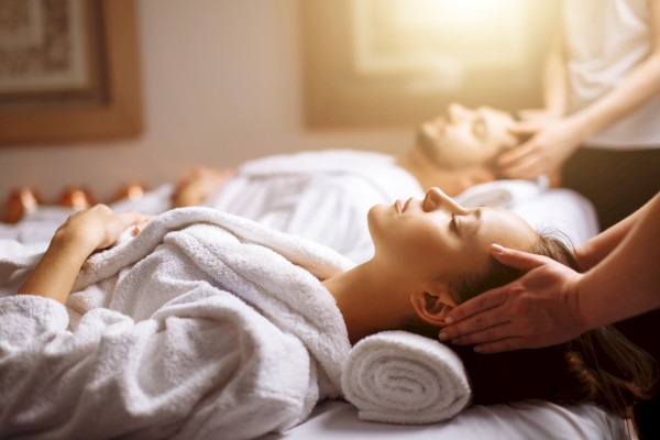 People relaxing on massage tables, wearing white robes, receiving head massages in a spa setting with warm lighting.