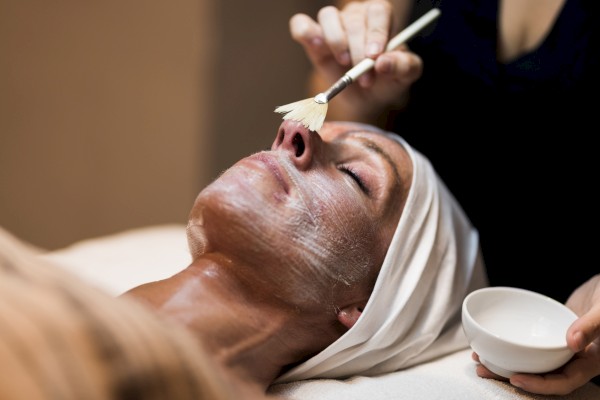 A person is receiving a facial treatment, lying down with a white cream mask applied using a brush, while holding a small bowl.