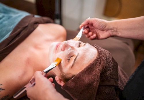 A person receives a facial treatment with a mask applied using brushes, lying down with a towel around their head.