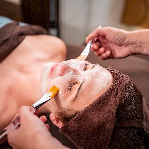 A person receives a facial treatment with a mask applied using brushes, lying down with a towel around their head.