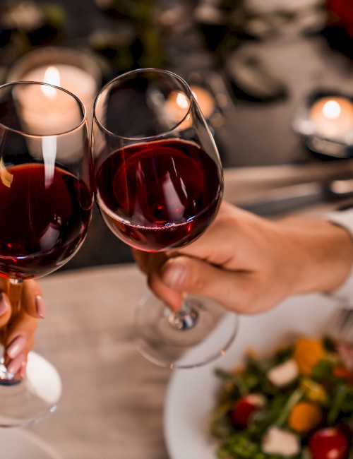 Two people clinking wine glasses over a candlelit dinner table with a salad and candles in the background.