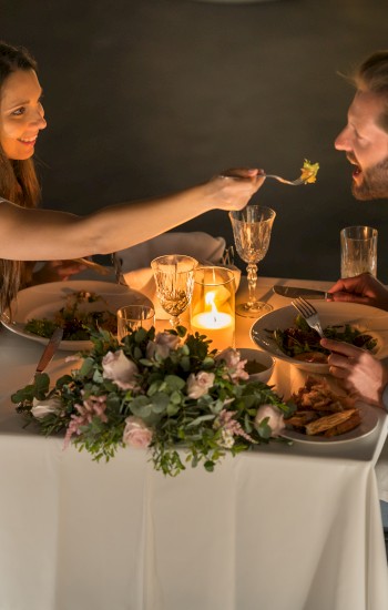 A couple enjoys a romantic candlelit dinner, with one person feeding the other at a beautifully decorated table, sharing a joyful moment.