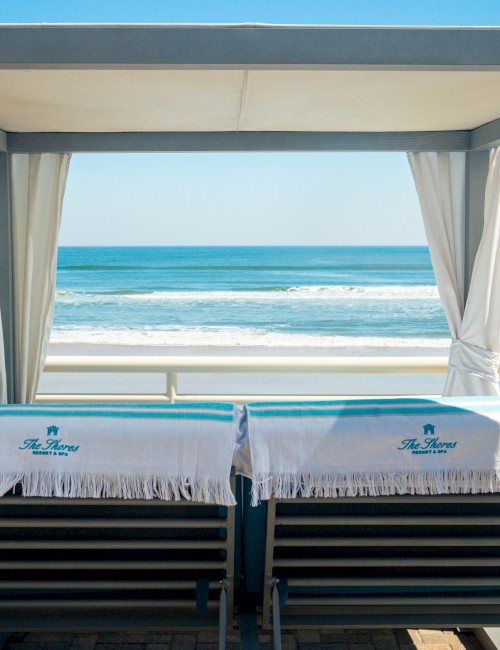 Two lounge chairs under a cabana face the ocean, with towels draped over them, offering a serene seaside view.