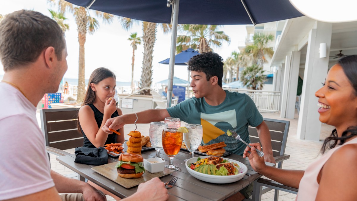 A group of people enjoy a meal together outdoors at a beachside restaurant, with drinks and various dishes on the table.