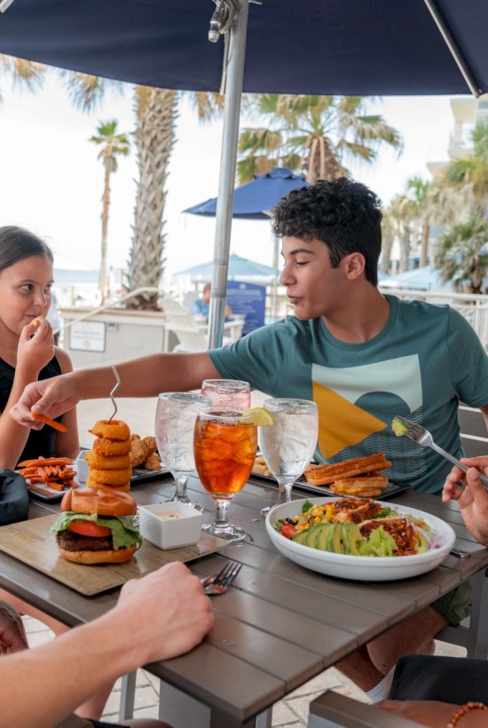A group of people enjoying a meal at an outdoor restaurant by the beach, with plates of food, drinks, and palm trees in the background.