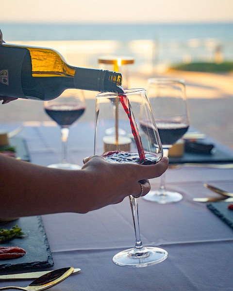 A person is pouring red wine into a glass on a table set with plates, with a scenic outdoor view in the background.