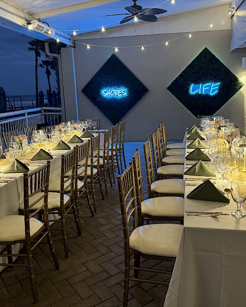 Elegant dining setup with long tables, white tablecloths, and green napkins; neon signs on the wall read 