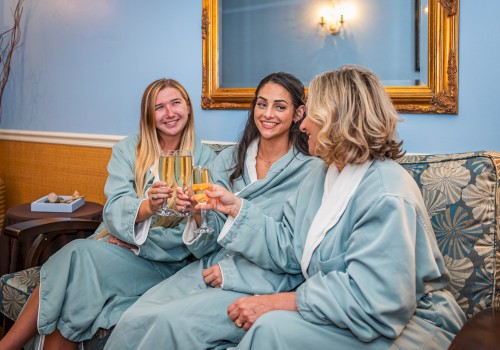 Three people in robes are sitting on a couch, toasting with drinks in a cozy room with a blue wall and a decorative mirror.