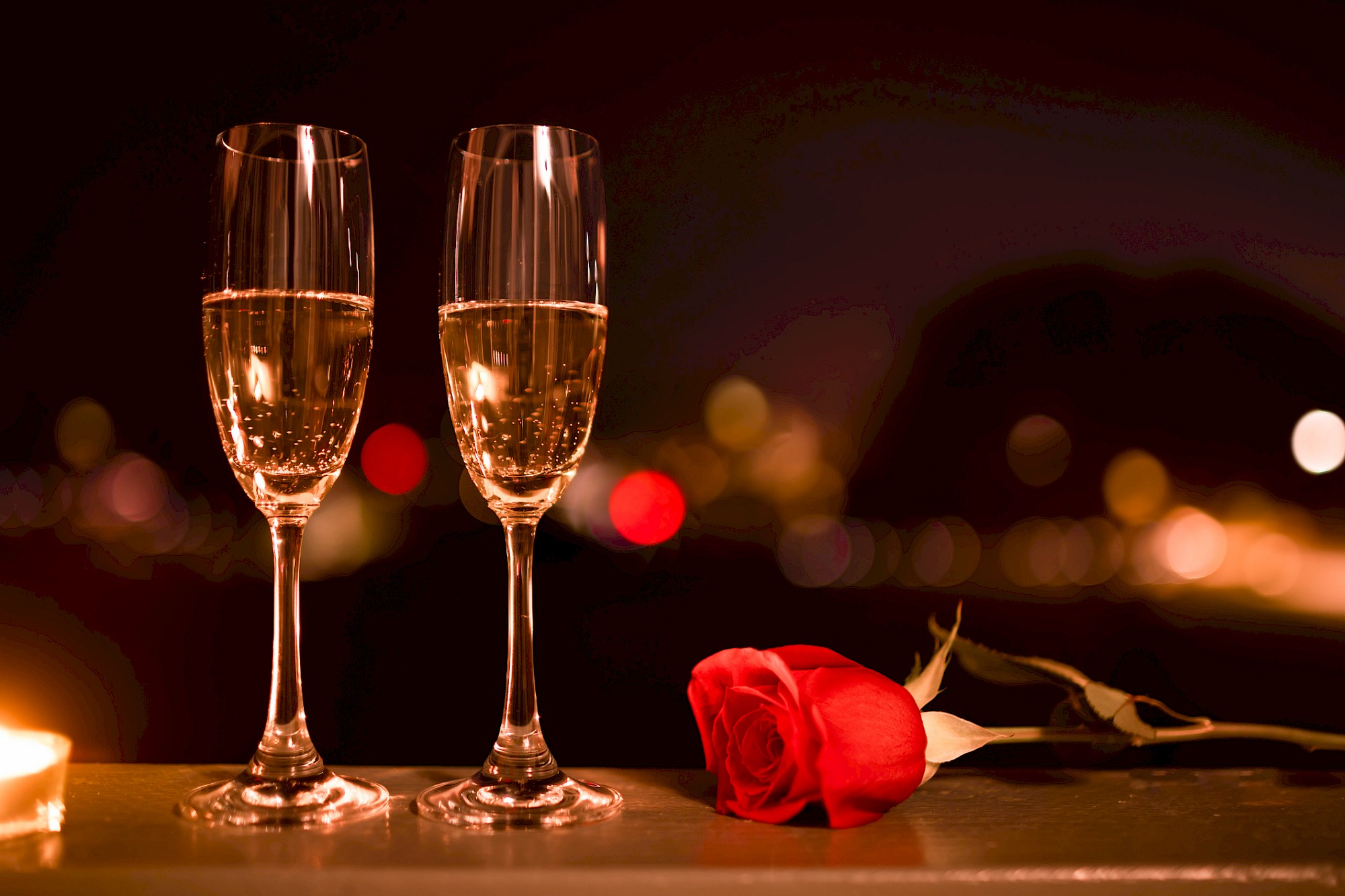 Two champagne flutes and a red rose on a table, with blurred colorful lights in the background, creating a romantic setting.