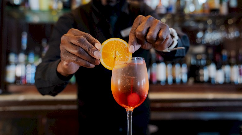 A bartender garnishing a cocktail with an orange slice in front of a bar filled with bottles.