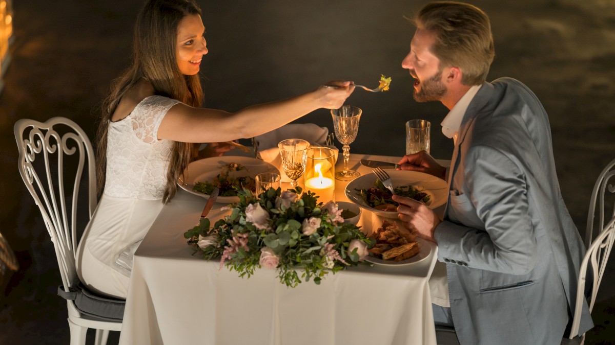 A couple enjoys a romantic candlelit dinner, with the woman feeding the man at a beautifully decorated table.